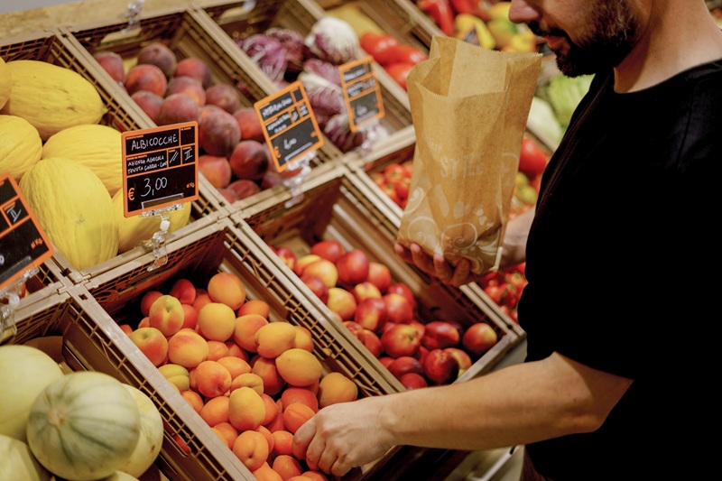 Uomo con sacchetto di carta sceglie frutta al banco di Buò, cartello «Albicocche € 3,00», meloni e susine in cassette