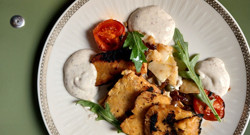 Tempeh grigliato con pomodori arrosto, rucola, capperi e salsa chiara su piatto bianco bordato d'oro, tavolo verde
