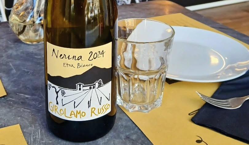 Bottiglia di Etna Bianco Nerina 2024 Girolamo Russo accanto a un bicchiere e un piatto bianco su tovaglietta gialla