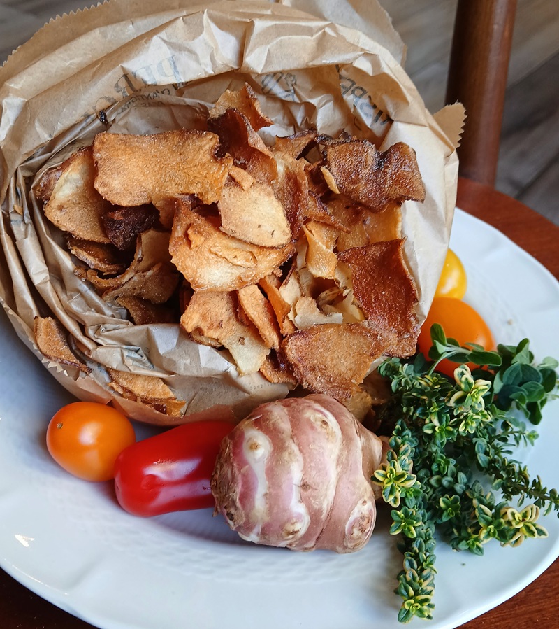 Chips di topinambur in un cartoccio di carta su piatto bianco con tubero crudo, pomodorini, peperoncino rosso e timo